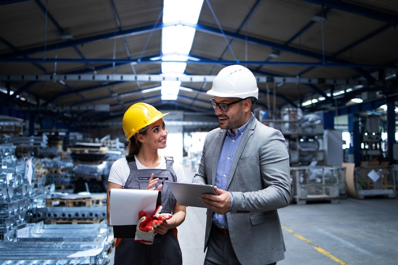 A man wearing a white hard hat and business suit reviews information on a tablet with a female worker in a yellow helmet and industrial uniform, inside a warehouse or production facility.