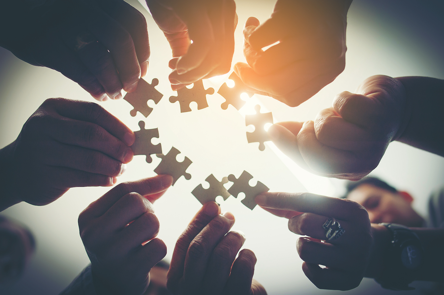 Several hands working together to connect puzzle pieces, symbolising teamwork, collaboration, and unified problem-solving under a warm light.