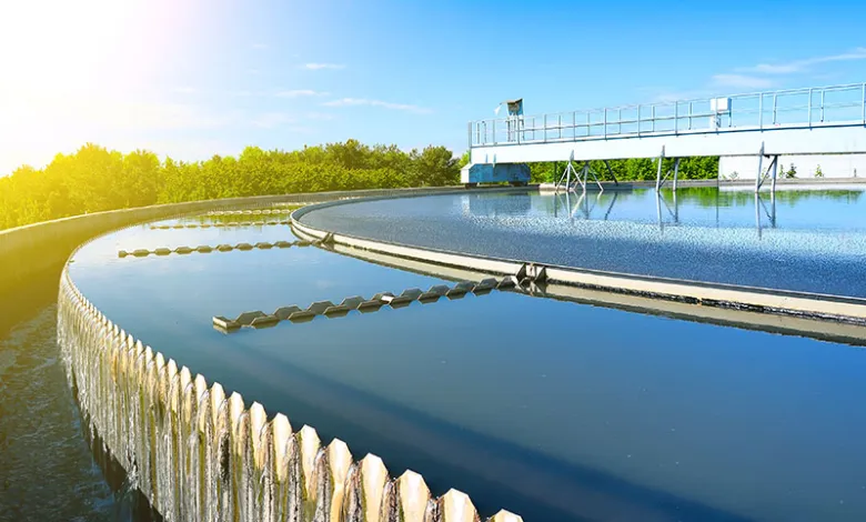 View of an outdoor industrial wastewater treatment plant. A large circular tank with clean, bright water overflows along serrated edges, surrounded by greenery and metal structures. The scene conveys advanced technology and respect for the environment in water management.