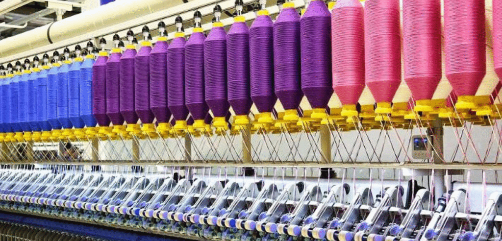 A textile production facility showing rows of colorful spools of thread in purple, blue, and pink, organized on a machine used in the manufacturing process.