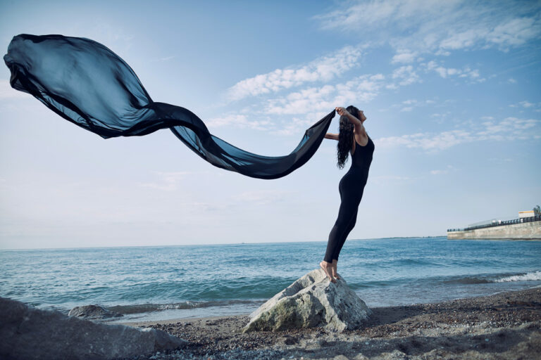 Mujer de pie sobre una roca en la playa, sosteniendo un trozo de tela negra que ondea al viento, con el mar y el cielo azul de fondo.