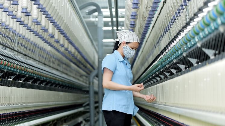 female textile worker wearing a face mask and headscarf inspects threads in a spinning mill.