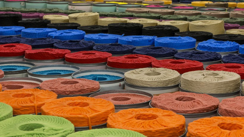 Spools of dyed textile fibres in a wide range of vibrant colours, neatly arranged in rows, ready for use in fabric manufacturing processes.
