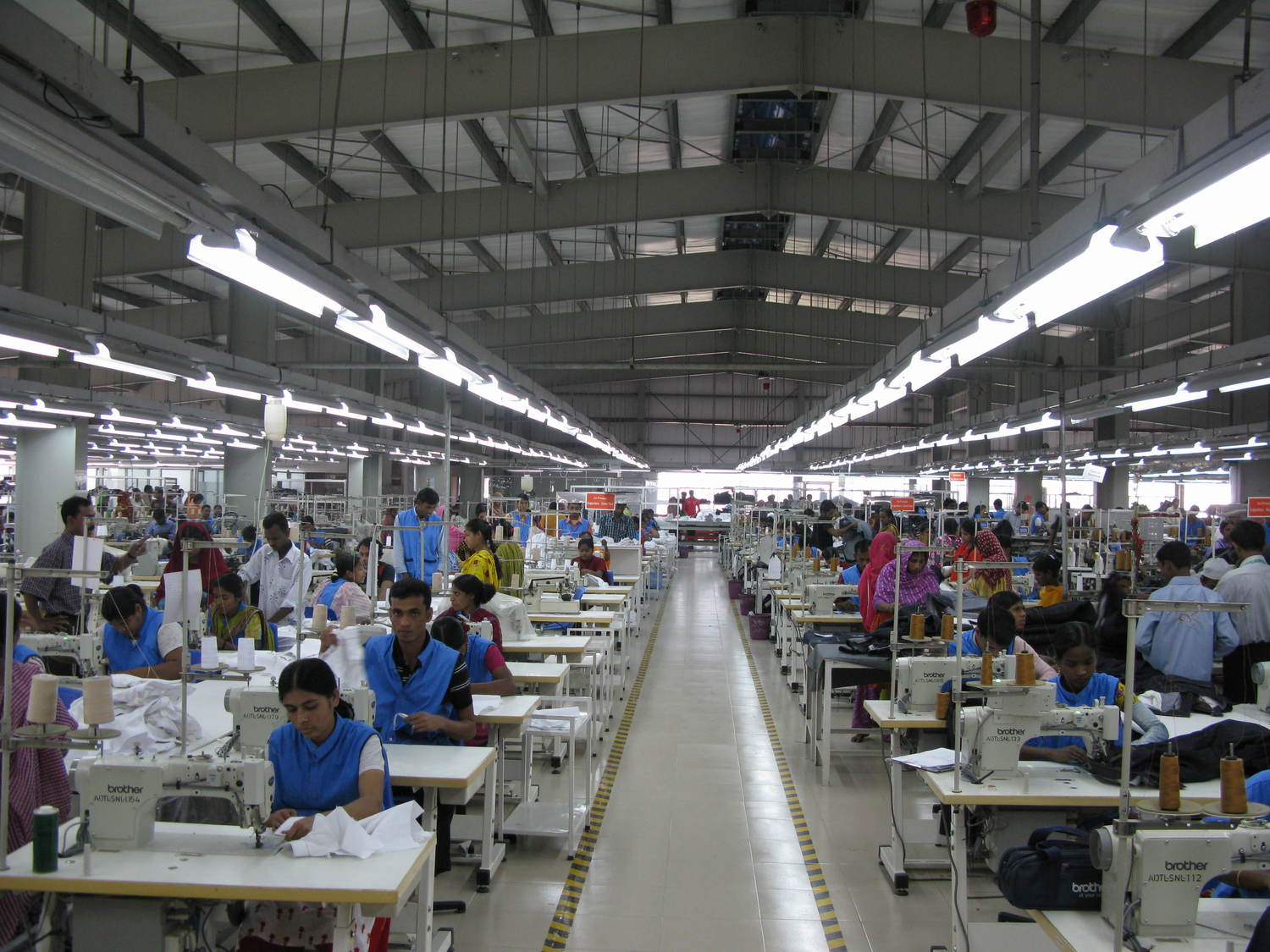 A spacious, modern textile factory with rows of workers operating sewing machines on either side of a wide central aisle, illustrating industrial-scale garment production.